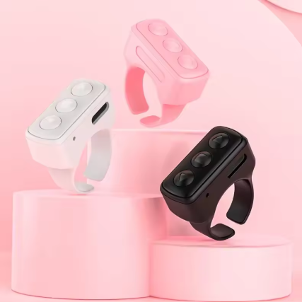 Three ring-shaped wearable remote control devices with rectangular control panels are displayed on pink pedestal platforms. The devices come in white, light pink, and black colorways, set against a soft pink background.