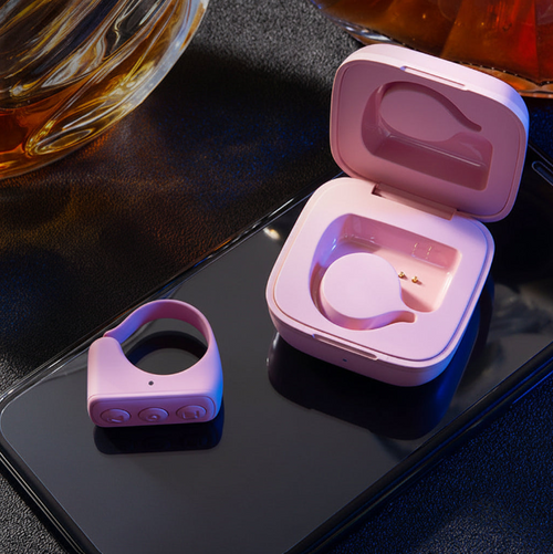 A pink wearable ring-shaped device with a matching open charging case sits on a dark smartphone screen, with a glass of amber liquid partially visible in the upper left.